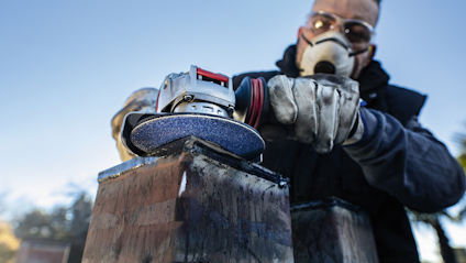 Person i skyddsglasögon och mask använder en vinkelslip på metall.