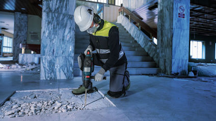 En person i säkerhetsutrustning använder en borrmaskin på ett betonggolv i en inomhusmiljö med marmorväggar och trappor.