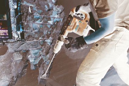 Person som använder en blå borrmaskin på en tegelvägg. Bär handskar och jeans.