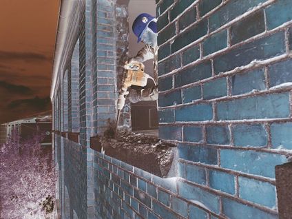 Person i gul hjälm borrar i röd tegelvägg med eldriven borrmaskin.