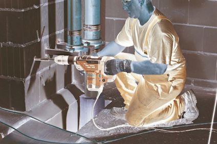 Person i arbetskläder borrar i en vägg med en stor blå borrmaskin. Rör och kablar syns i omgivningen.