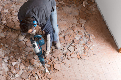 En person använder en borrhammare på ett golv täckt av krossat kakel och damm.