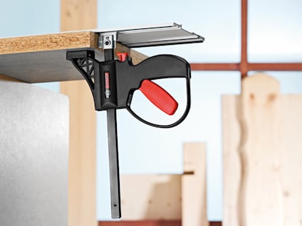 Limklämma applicerad på en bräda, med röd och svart handtag i en verkstadsmiljö.
