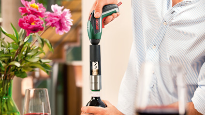 En person använder en elektrisk korkskruv för att öppna en vinflaska, blommor och vinglas i bakgrunden.