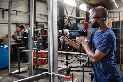 Man i verkstad arbetar med ett elverktyg på stålkonstruktion. Bakgrund med industriell utrustning och verktyg.