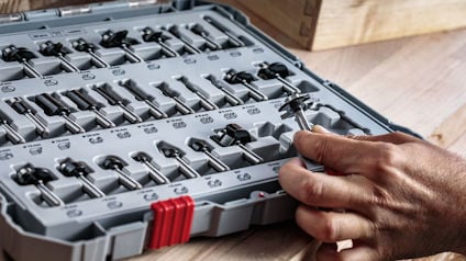 Öppen låda med olika borrbits. En hand håller i en bit. Bitarna är organiserade i rader i lådan.