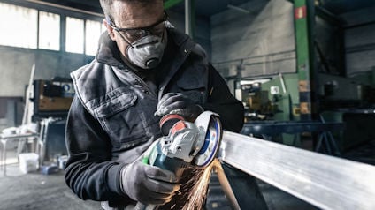 Person slipar metall med skyddsutrustning och vinkelslip i en verkstad.