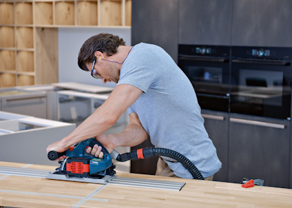 Person använder en cirkelsåg på en träyta i ett kök, med hörselskydd och skyddsglasögon.