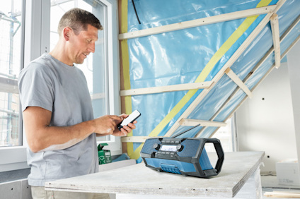 Man i grå tröja vid fönster, använder smartphone. På bordet står en blå ljudanläggning. Byggmaterial i bakgrunden.