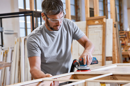 Person slipar träbit med elektrisk slipmaskin i en verkstad. Bär skyddsglasögon och hörselskydd.