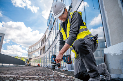 En person i hjälm och säkerhetsväst använder en elektrisk borr på en byggarbetsplats.