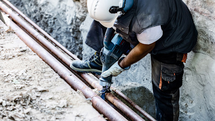 Person bär skyddshjälm och borrmaskin, arbetar på rör i grävd grop.