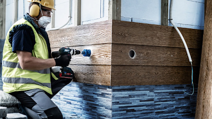 Person i skyddshjälm och väst använder borrmaskin vid panel av trä och kakel.