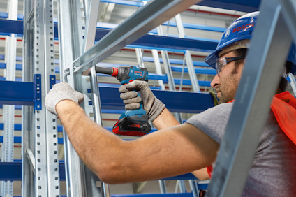 En man använder en blå sladdlös borrmaskin för att montera metallramar, iklädd skyddshjälm och handskar.