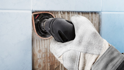 Handskar klädd hand håller en elverktyg mot kaklat vägg, avlägsnar gammalt fogmaterial.