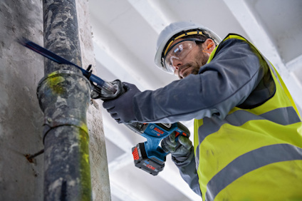 En person i hjälm och gul väst använder en blå borrmaskin på ett rör inomhus.