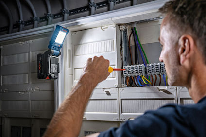 Person arbetar i elcentral med skruvmejsel, omgiven av kablar. Belysning från lampa ovanför.