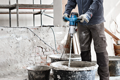Person blandar material i en hink med en elektrisk mixer, omgiven av byggarbetsplats.