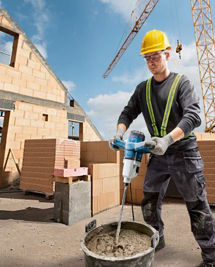 Byggarbetare blandar cement i en hink med en elektrisk mixer; bakom syns en byggnad under uppförande och en kran.