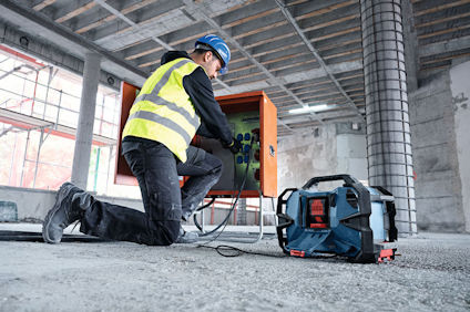 Person knäböjer i en byggarbetsplats och arbetar med elektrisk utrustning; bär hjälm och reflexväst. Verktygslåda syns närmast.