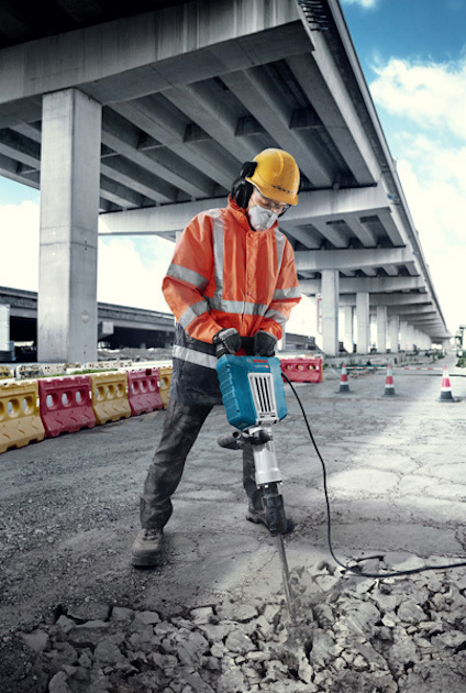 Arbetare i orange skyddsutrustning använder en tryckluftsborr under en bro på en grusväg.
