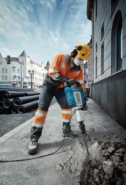 Arbetare i orange skyddskläder och hjälm använder en bilningsmaskin på trottoar. Rör ligger staplade i bakgrunden.