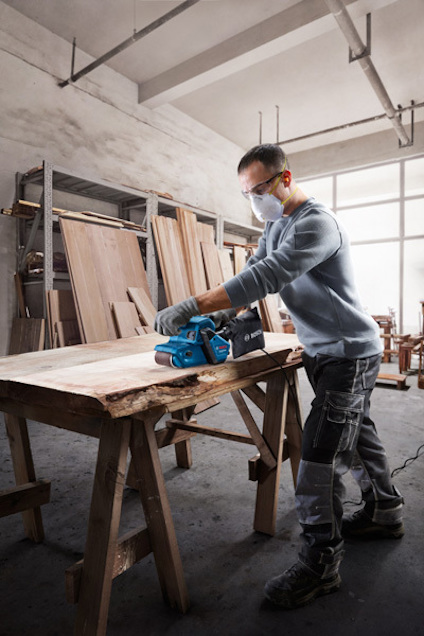 En person i skyddsutrustning använder en blå bandslip på en träskiva i en verkstad.