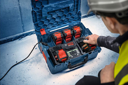 Öppen verktygslåda med laddare och tre batterier. En person i hjälm och väst håller kabel.