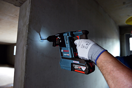 En person borrar i en betongvägg med en Bosch 18V borrmaskin, iklädd arbetshandskar och hjälm.