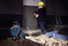 En person i hjälm arbetar med en power tool vid betongstruktur i byggmiljö, med skyddsutrustning i närheten.