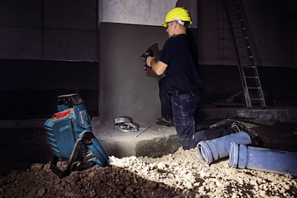En person i hjälm arbetar med en power tool vid betongstruktur i byggmiljö, med skyddsutrustning i närheten.