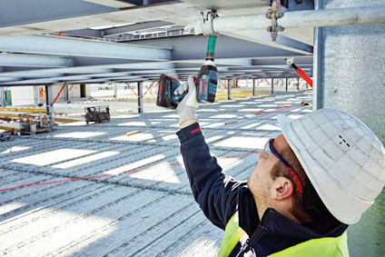 Person med hjälm och skyddsglasögon använder en borrmaskin för att fästa metallbalkar.