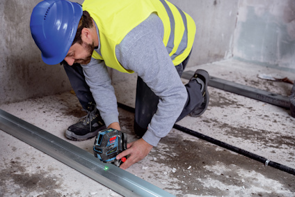 Person med blå hjälm och gul väst använder en grön laserverktyg på betonggolv.