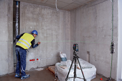 Byggnadsarbetare i hjälm och väst markerar på betongvägg med linjelaser och stativ bredvid täckt materialbunt.