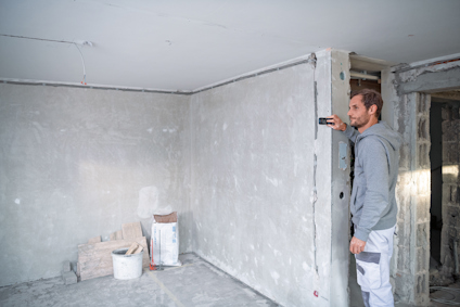 En man mäter vägg med verktyg i ett rum under renovering. Byggmaterial och tråg synliga på golvet.