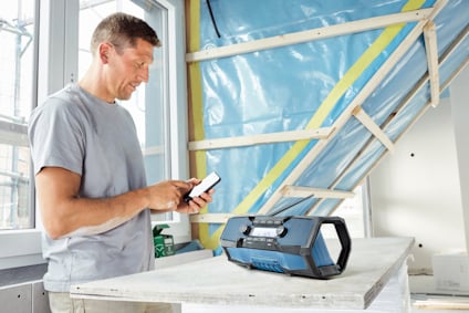 Man in a t-shirt using a smartphone next to a portable radio on a wooden workbench in a partially constructed room.