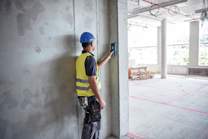 Person med blå hjälm och gul väst använder en elektronisk mätare på en betongvägg i en byggarbetsplatsmiljö.