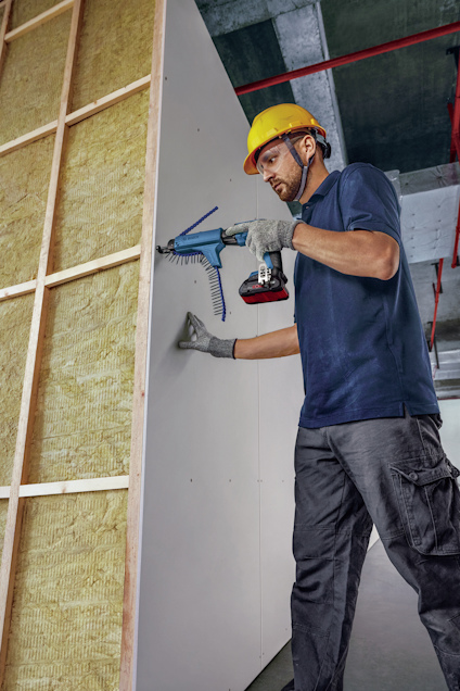 Person i arbetshjälm monterar gipsskiva på väggstomme med en skruvautomat i ett byggprojekt.