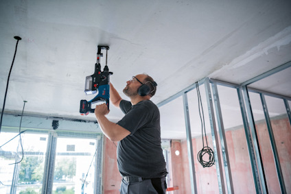 Man using a power tool on a ceiling in a room under construction, wearing protective headphones.