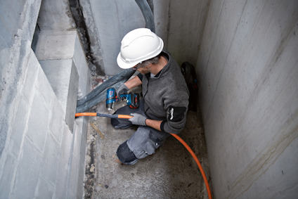 Person i byggkläder och hjälm använder en borrmaskin på en betongvägg nära ett hörn, orange kabel syns.
