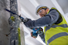 Person i byggarbetskläder använder en blå och röd elektrisk såg på två metallrör mot en betongvägg.