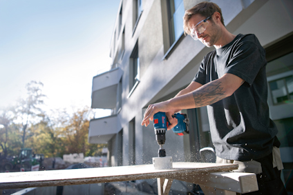 Person borrar hål i träskiva med blå och röd borrmaskin. Skyddsglasögon och svart t-shirt.