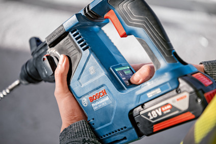 Närbild av händer som håller i en blå Bosch Professional 18V borrmaskin med borr på grå betongyta.