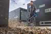 Man in gray clothing using a blue leaf blower on fallen leaves outside modern black building.