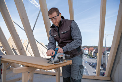 En person i skyddsglasögon sågar en träplanka med en elektrisk såg på en byggarbetsplats.