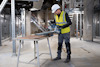 Person med skyddshjälm och gul väst använder verktyg i industriell miljö.