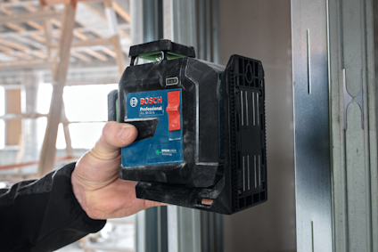 En person håller en Bosch Professional lasermätare framför en stålregel i en byggnad under konstruktion.
