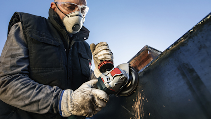 En person använder en vinkelslip på en metallplatta, med skyddsmask och handskar.