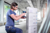Man in a workshop using a power drill on a metal cabinet, wearing safety glasses and a blue shirt.