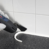 Hand använder ett elverktyg med spackelblad för att avlägsna tätningsmassa från en kaklad vägg ovanför en arbetsyta.
